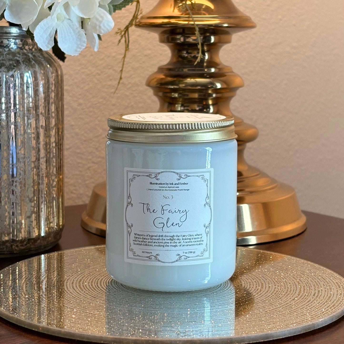 The Fairy Glen No. 3 wild heather and pine scented candle by Illumination by Ink and Ember, displayed on a gold tray with brass lamp and florals