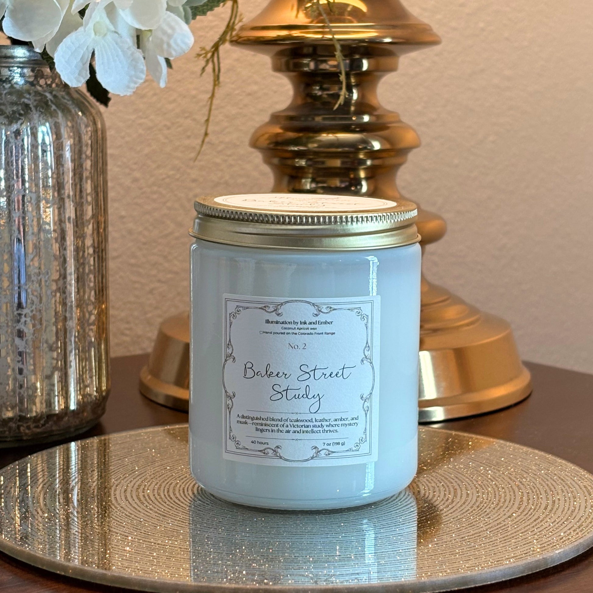Baker Street Study No. 2 teakwood, leather and amber scented candle by Illumination by Ink and Ember displayed on a gold tray with brass lamp and florals