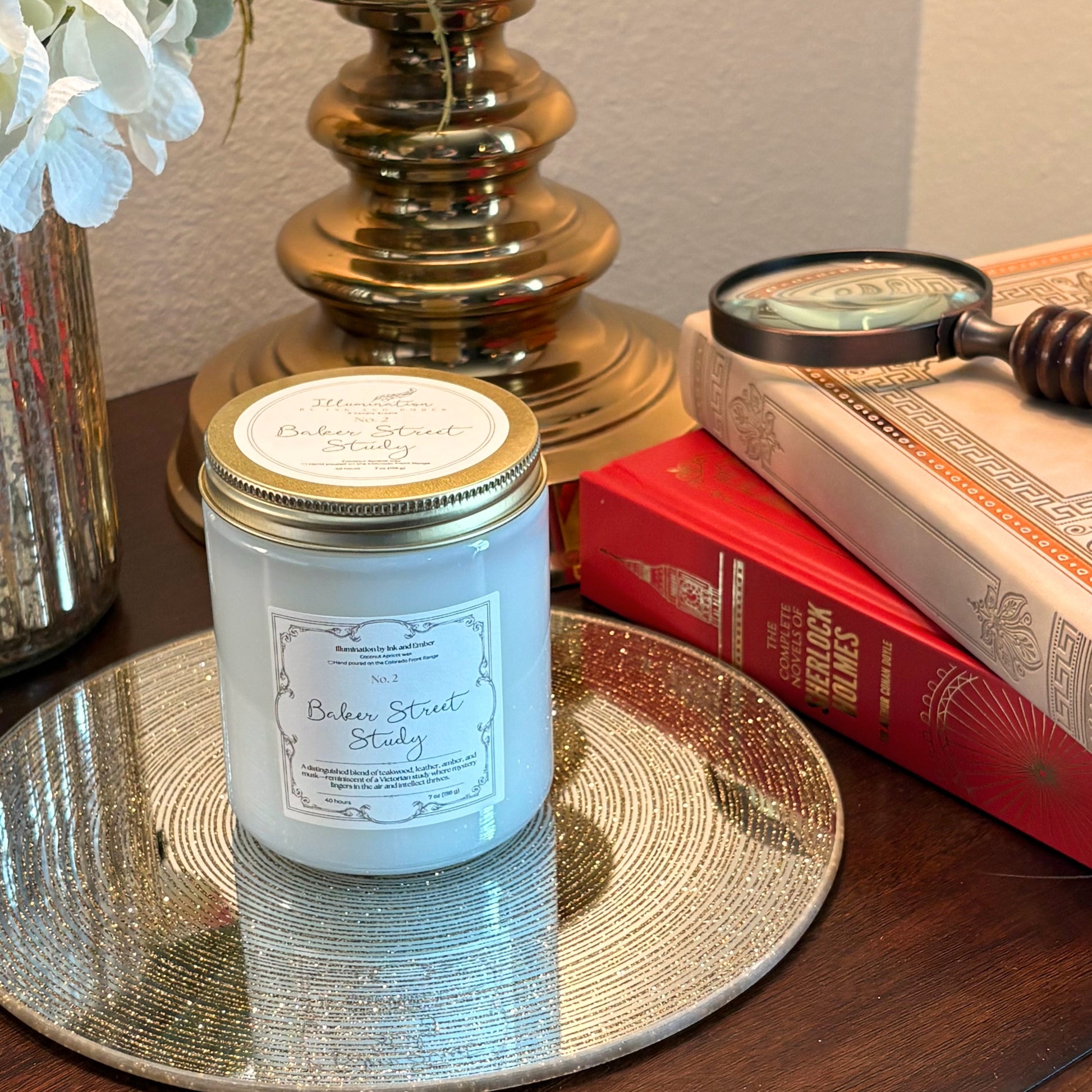 Baker Street Study No. 2 teakwood, leather and amber scented candle by Illumination by Ink and Ember, displayed on a gold tray beside Sherlock Holmes books and a vintage magnifying glass