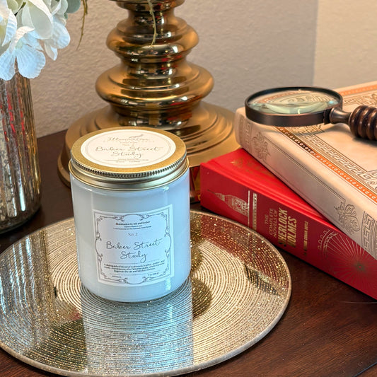 Baker Street Study No. 2 teakwood, leather and amber scented candle by Illumination by Ink and Ember, displayed on a gold tray beside Sherlock Holmes books and a vintage magnifying glass