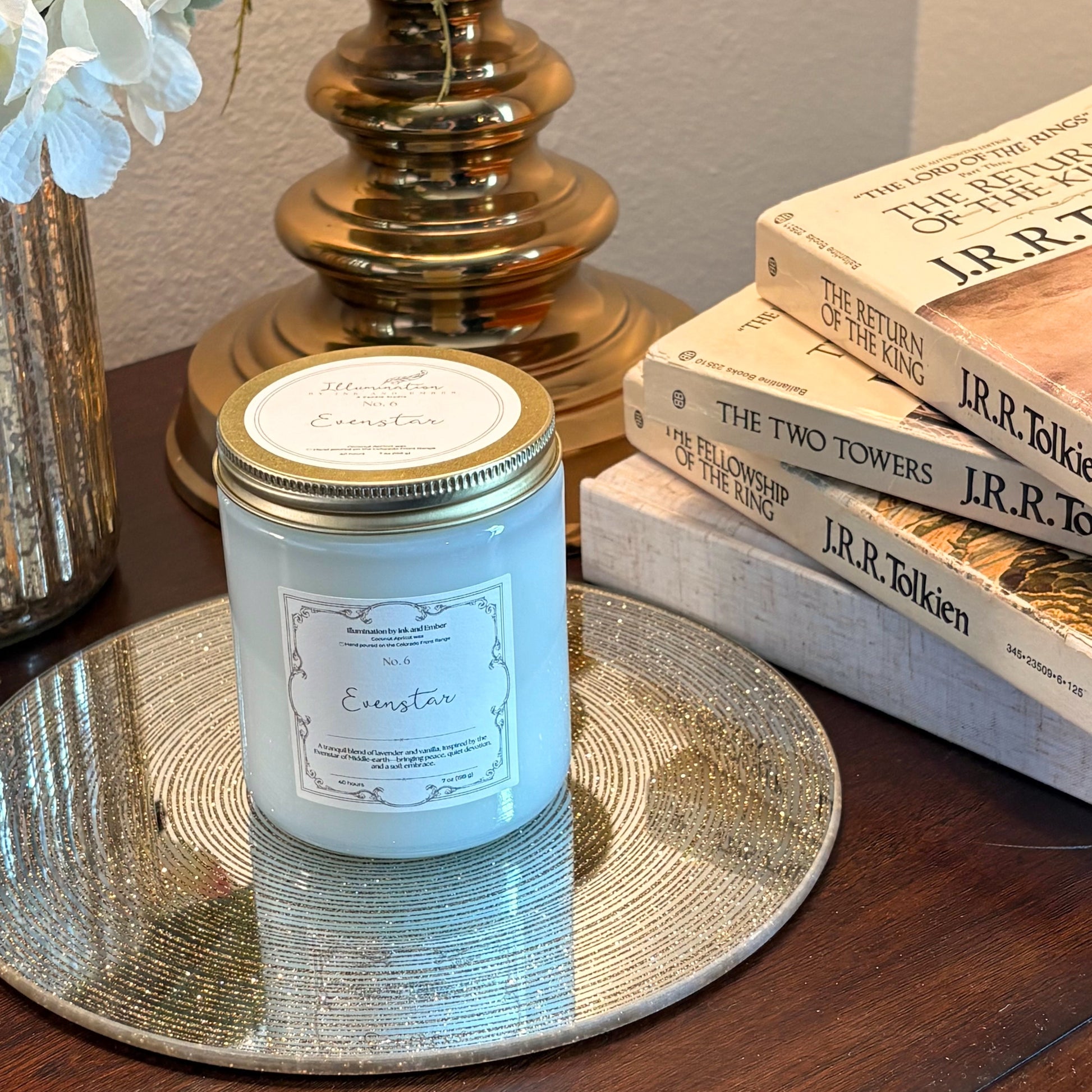 Evenstar No. 6 scented candle by Illumination by Ink and Ember, displayed on a gold tray beside a stack of J.R.R. Tolkien Lord of the Rings books