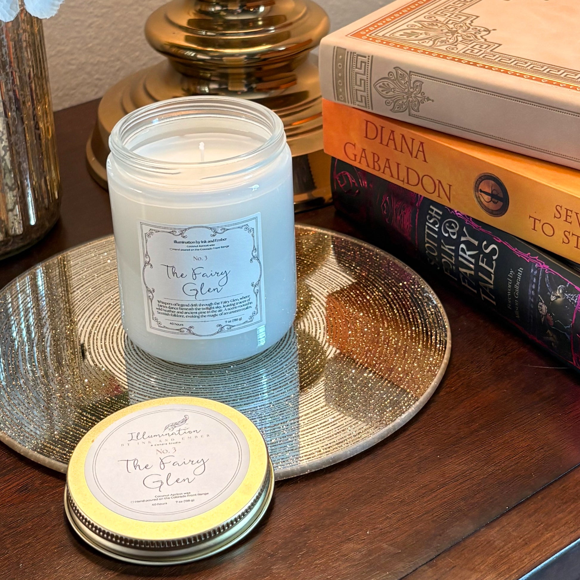 The Fairy Glen No. 3 wild heather and pine scented candle by Illumination by Ink and Ember, displayed on a gold tray beside Scottish Folk and Fairy Tales and Diana Gabaldon books with lid shown separately