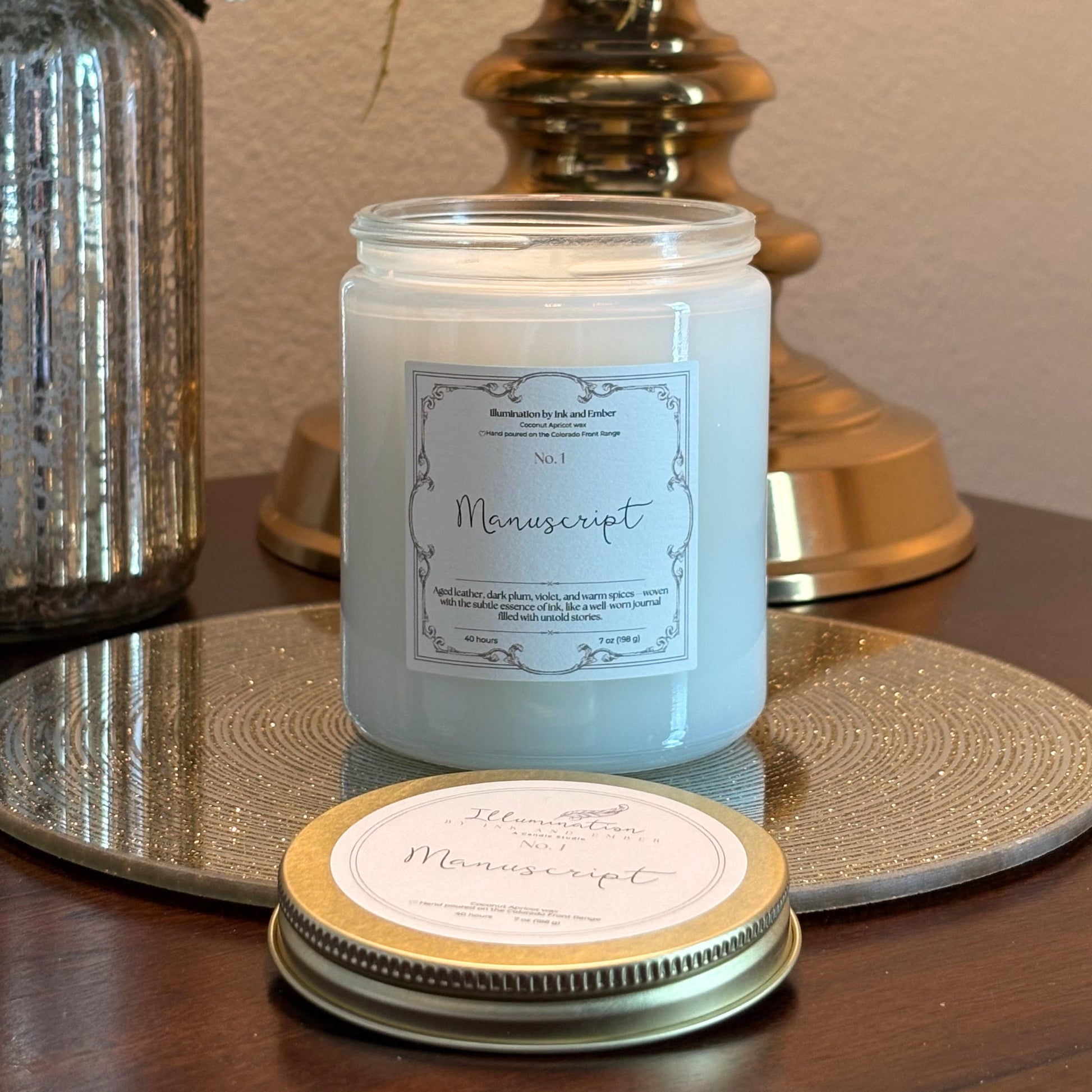 Manuscript No. 1 scented candle by Illumination by Ink and Ember displayed on a gold tray with lid shown separately, aged leather, dark plum, violet and warm spice fragrance