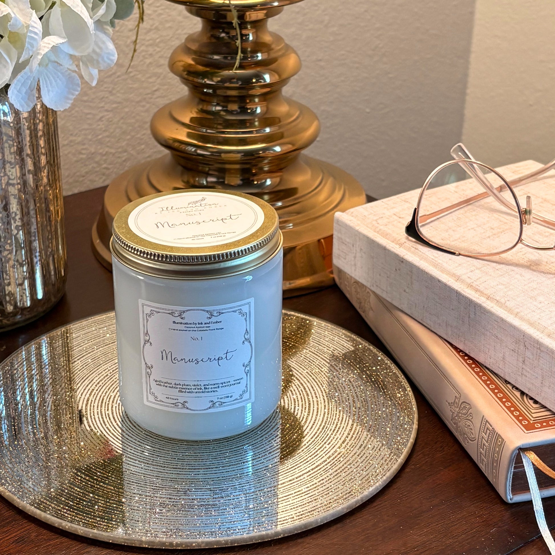 Manuscript No. 1 aged leather, dark plum and warm spice scented candle by Illumination by Ink and Ember on a gold tray beside stacked books and reading glasses