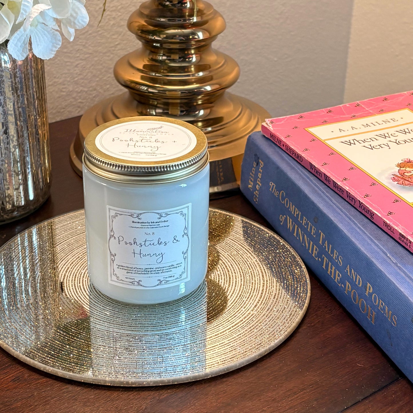 Poohsticks and Hunny No. 8 honey, jasmine and warm vanilla scented candle by Illumination by Ink and Ember displayed on a gold tray beside Complete Tales of Winnie-the-Pooh and A.A. Milne books