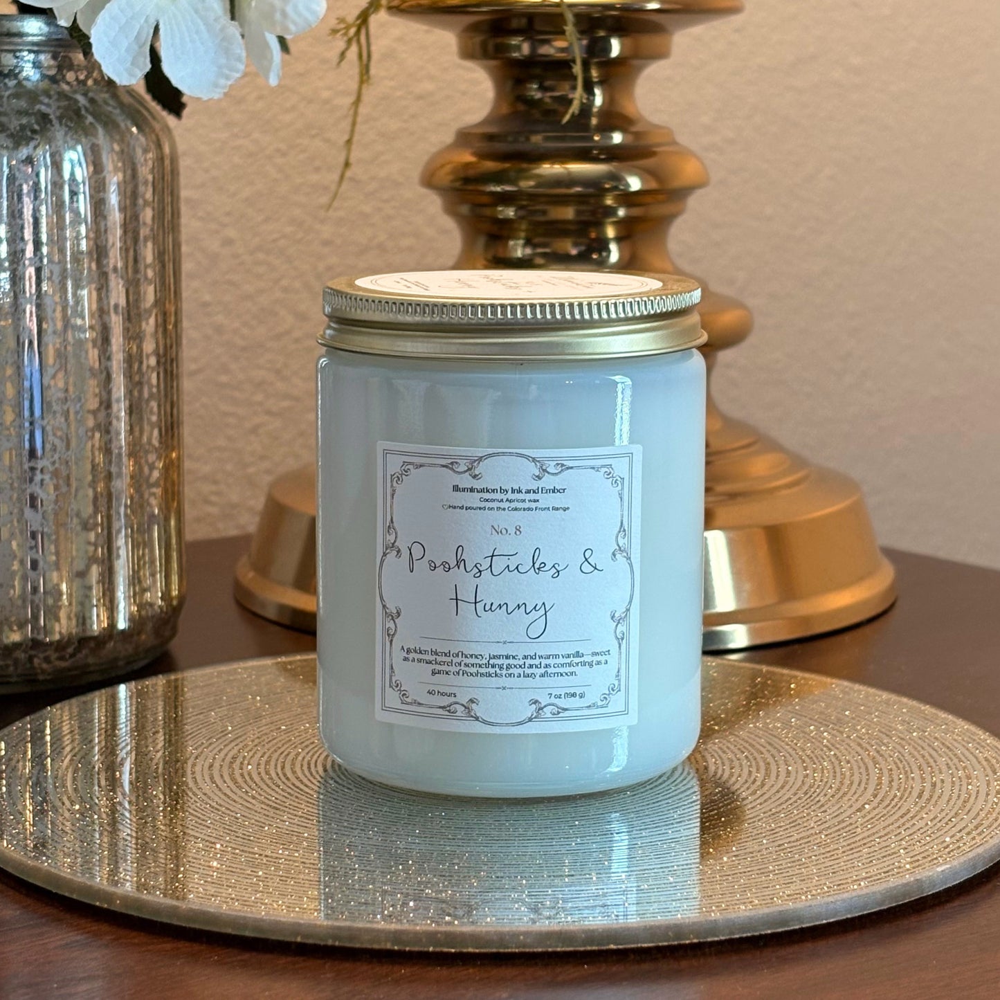 Poohsticks and Hunny No. 8 honey, jasmine and warm vanilla scented candle by Illumination by Ink and Ember displayed on a gold tray with brass lamp and florals