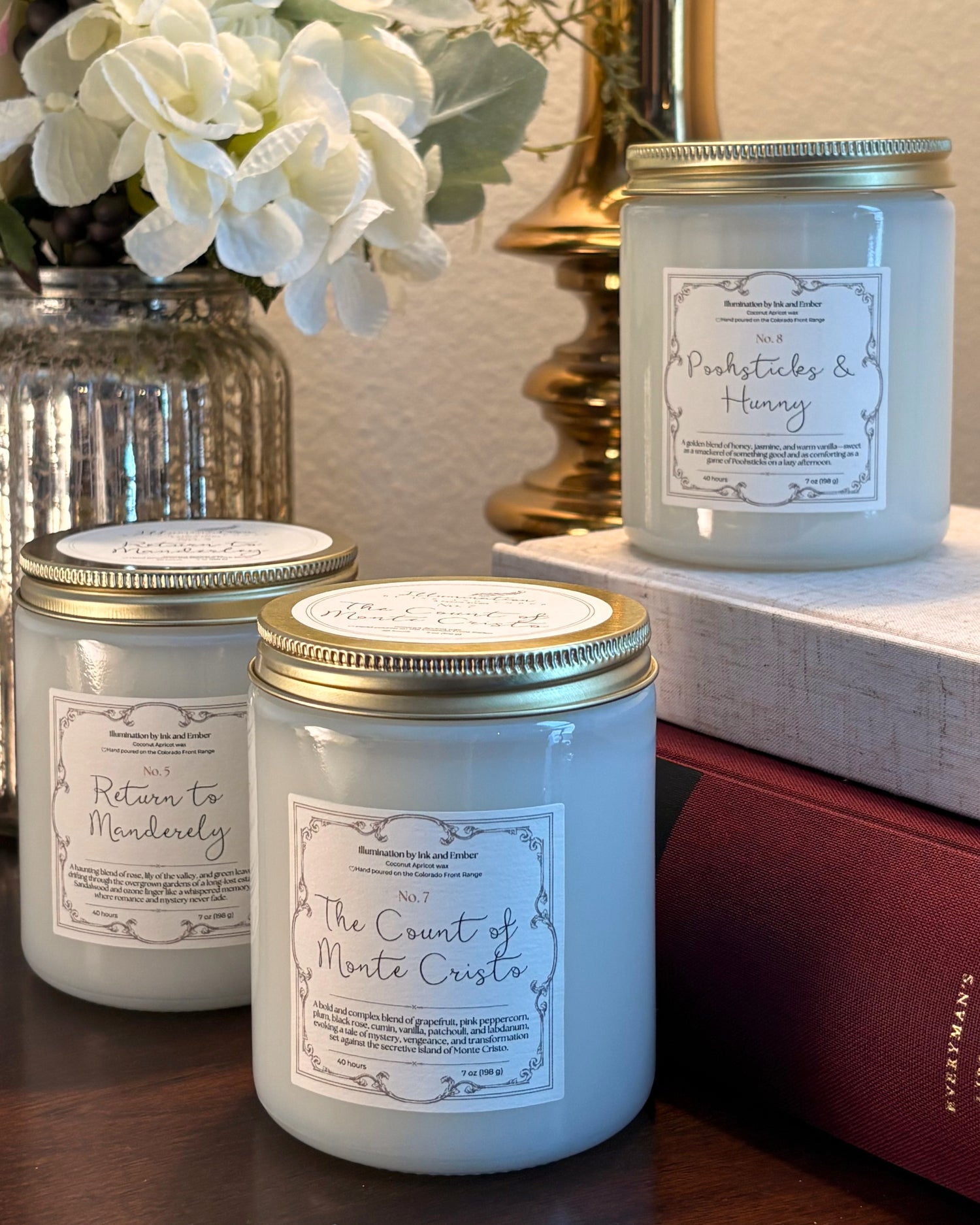 Illumination by Ink and Ember literary candle collection featuring Return to Manderley No. 5, The Count of Monte Cristo No. 7, and Poohsticks and Hunny No. 8 scented candles displayed with books and florals