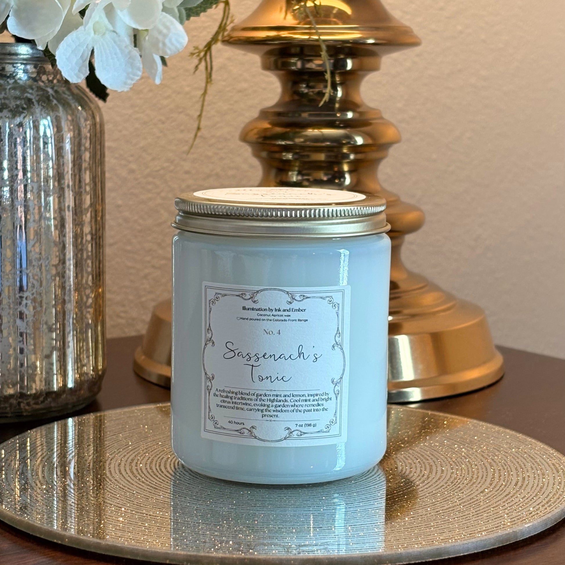 Sassenach's Tonic No. 4 garden mint and lemon scented candle by Illumination by Ink and Ember, displayed on a gold tray with brass lamp and florals
