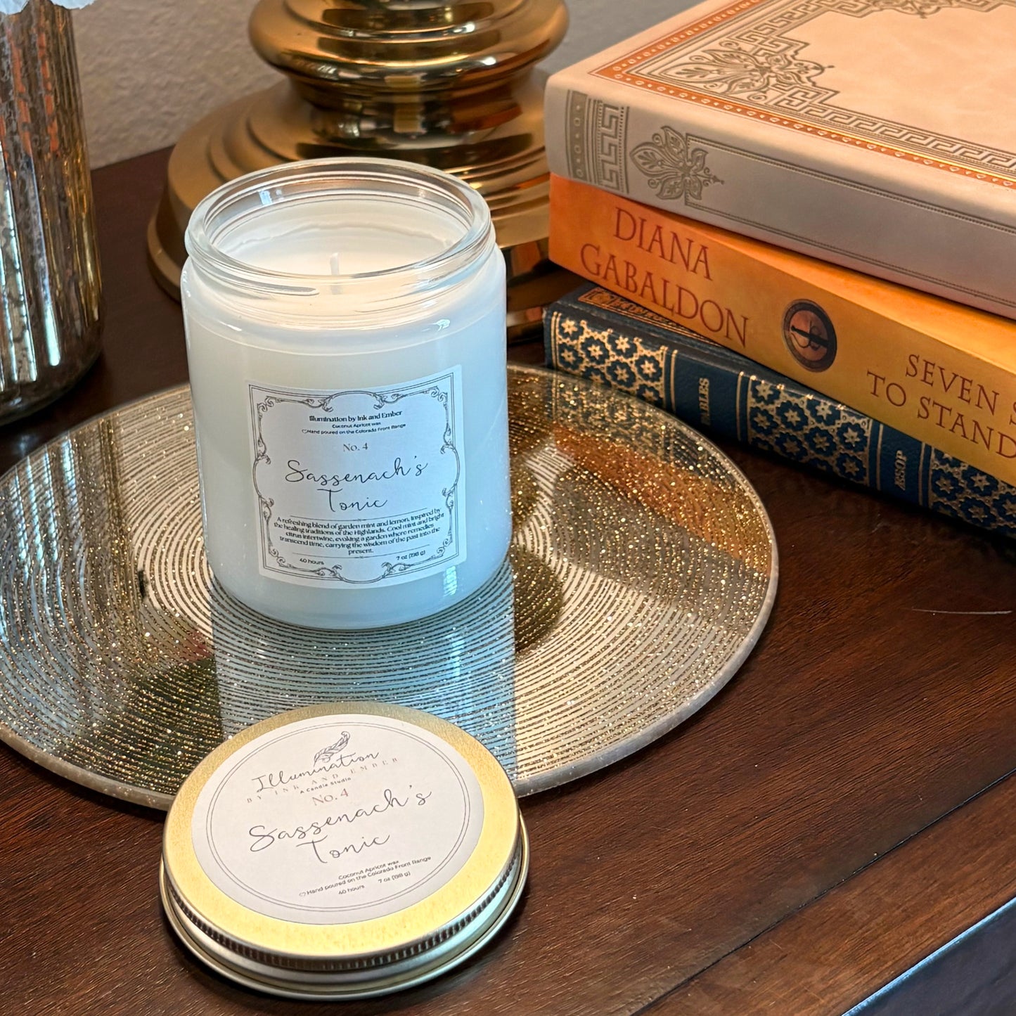 Sassenach's Tonic No. 4 garden mint and lemon scented candle by Illumination by Ink and Ember, displayed on a gold tray beside Diana Gabaldon Outlander books with lid shown separately